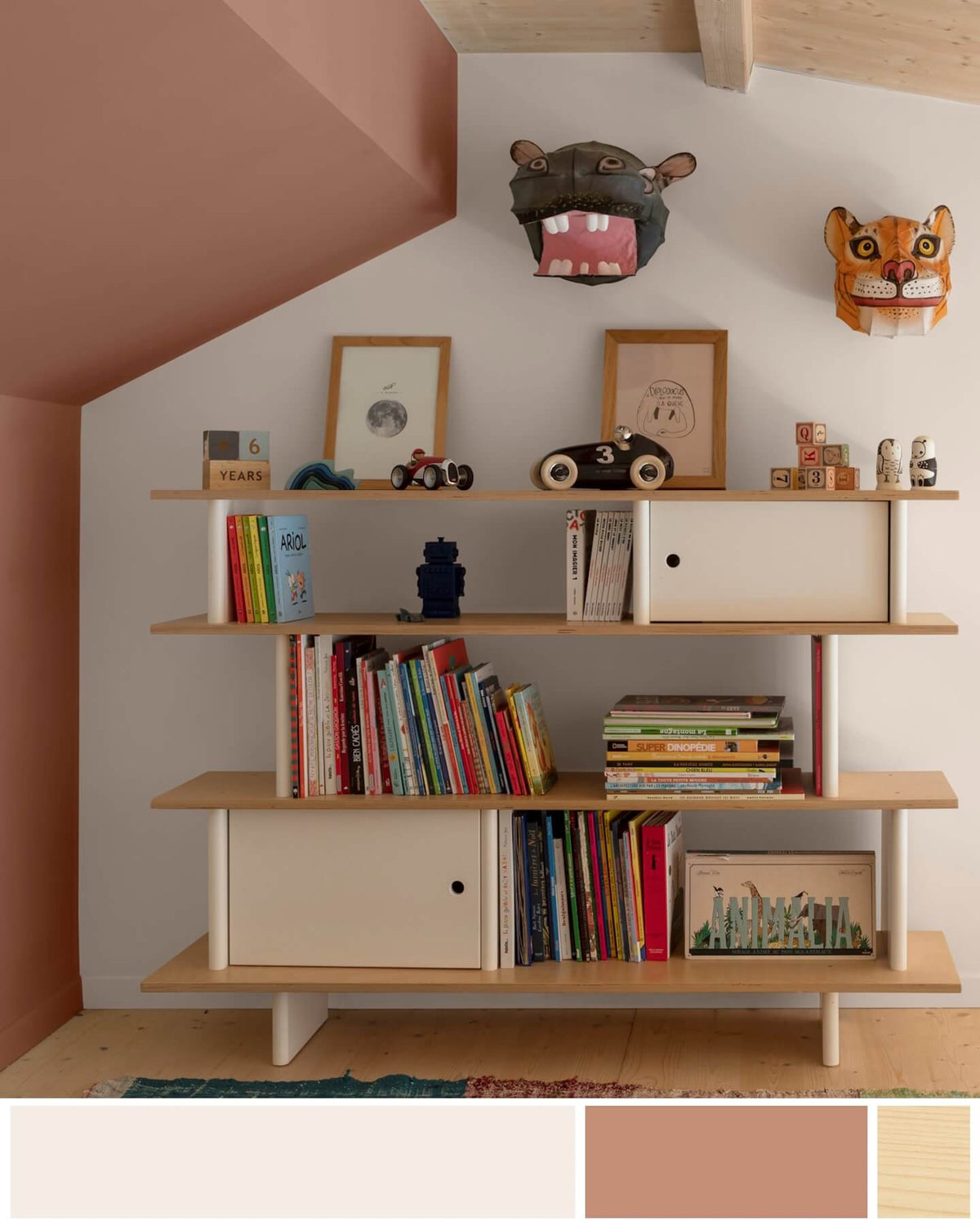 Chambre d'enfant sous les combles avec une grande bibliothèque en bois garnie de livres et jouets, des sculptures d'animaux accrochées, des murs peints avec la peinture Libéron 163 Faubourg, teintes Blanc Ballerine et Pêche Cancan, et un plafond lasuré au Badigeon teinte Bois Brut.