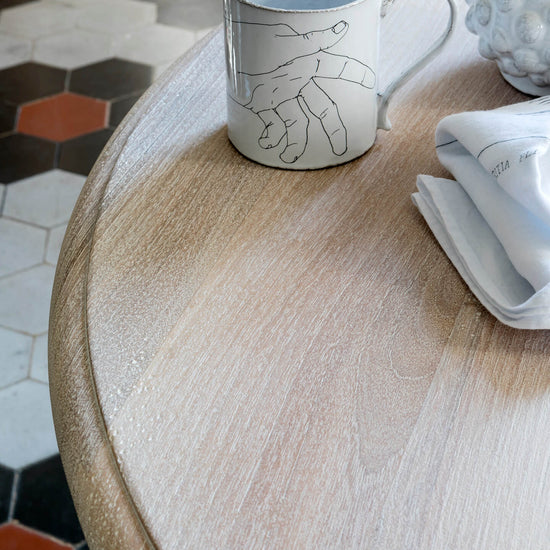 Table ronde en bois cérusé blanc Libéron, avec tasse décorée, linge clair et sol en carreaux graphiques.