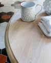 Table ronde en bois cérusé blanc Libéron, avec tasse décorée, linge clair et sol en carreaux graphiques.