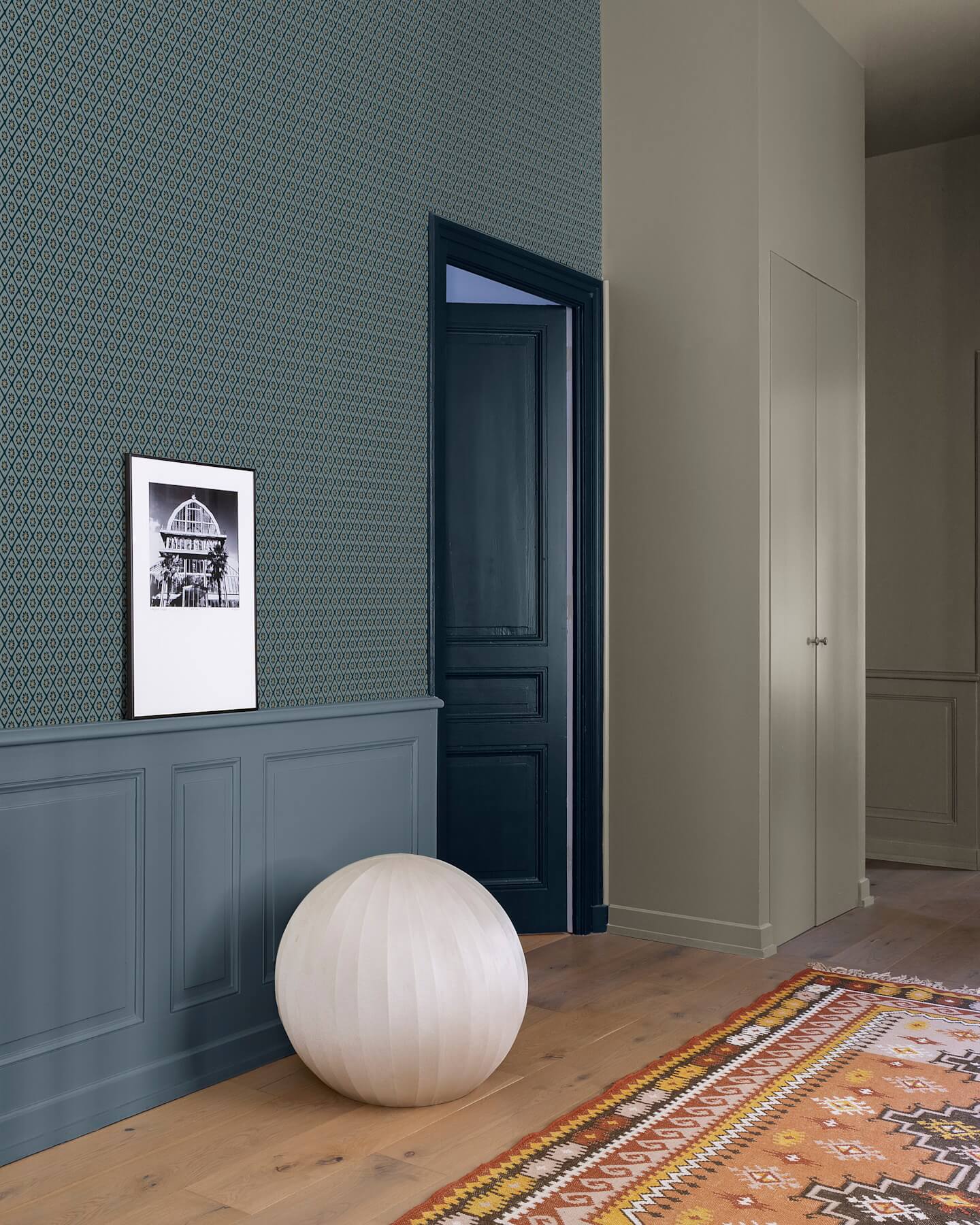 Couloir aux murs bleu grisé et papier peint graphique Libéron Maître Domino, avec boiseries et tapis coloré.