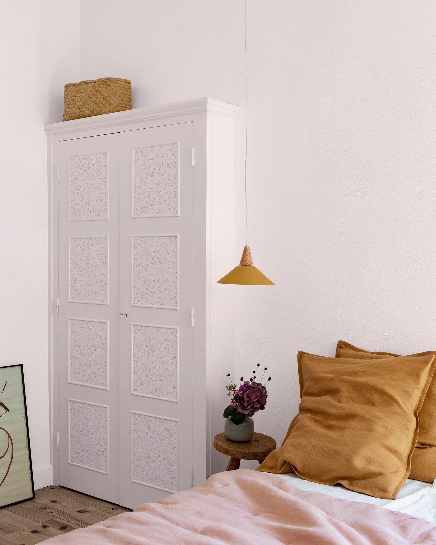 Chambre aux murs rose poudré, avec armoire blanche à panneaux floraux Belle Jeanne Libéron, suspension ocre et linge de lit rose.