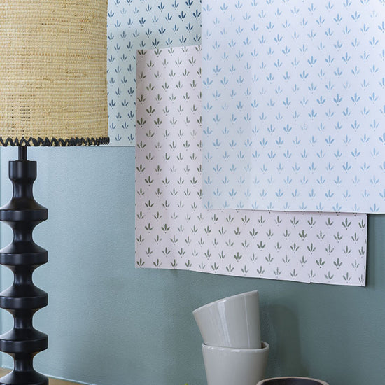 Échantillons des trois teinte du papier peint Velours de Papier Libéron accrochés sur un mur bleu, commode en bois avec lampe noire et beige.