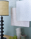 Échantillons des trois teinte du papier peint Velours de Papier Libéron accrochés sur un mur bleu, commode en bois avec lampe noire et beige.