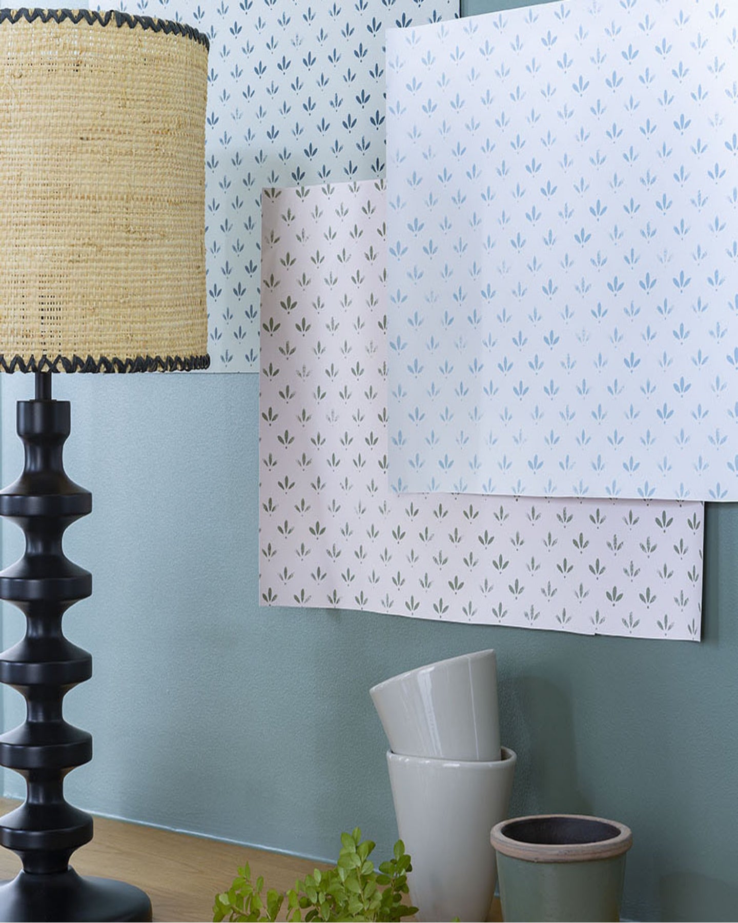Échantillons des trois teinte du papier peint Velours de Papier Libéron accrochés sur un mur bleu, commode en bois avec lampe noire et beige.