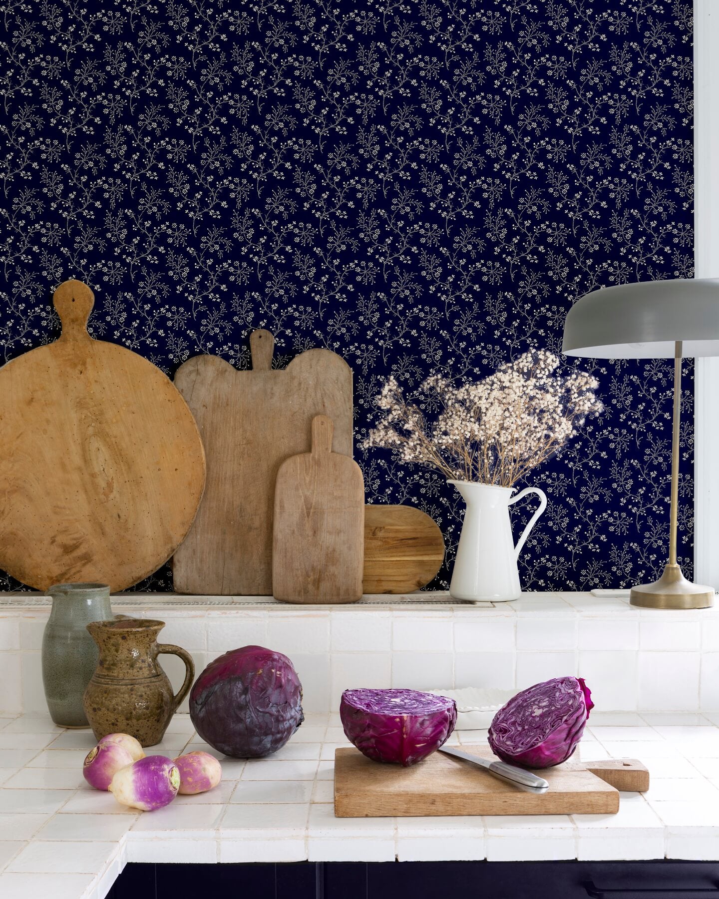 Cuisine au mur floral bleu nuit, teinte Belle Jeanne Libéron, avec plan de travail blanc, lampes et légumes violets.