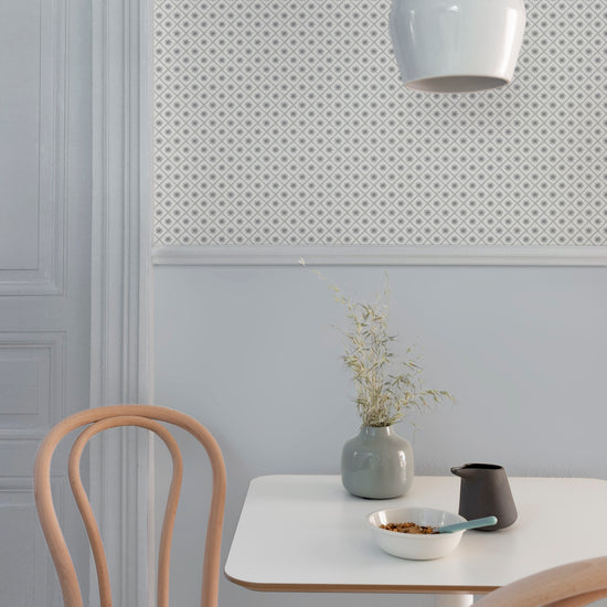 Coin repas aux murs bleu gris et motif géométrique Maître Domino Libéron, avec table blanche, chaises en bois clair et vase.