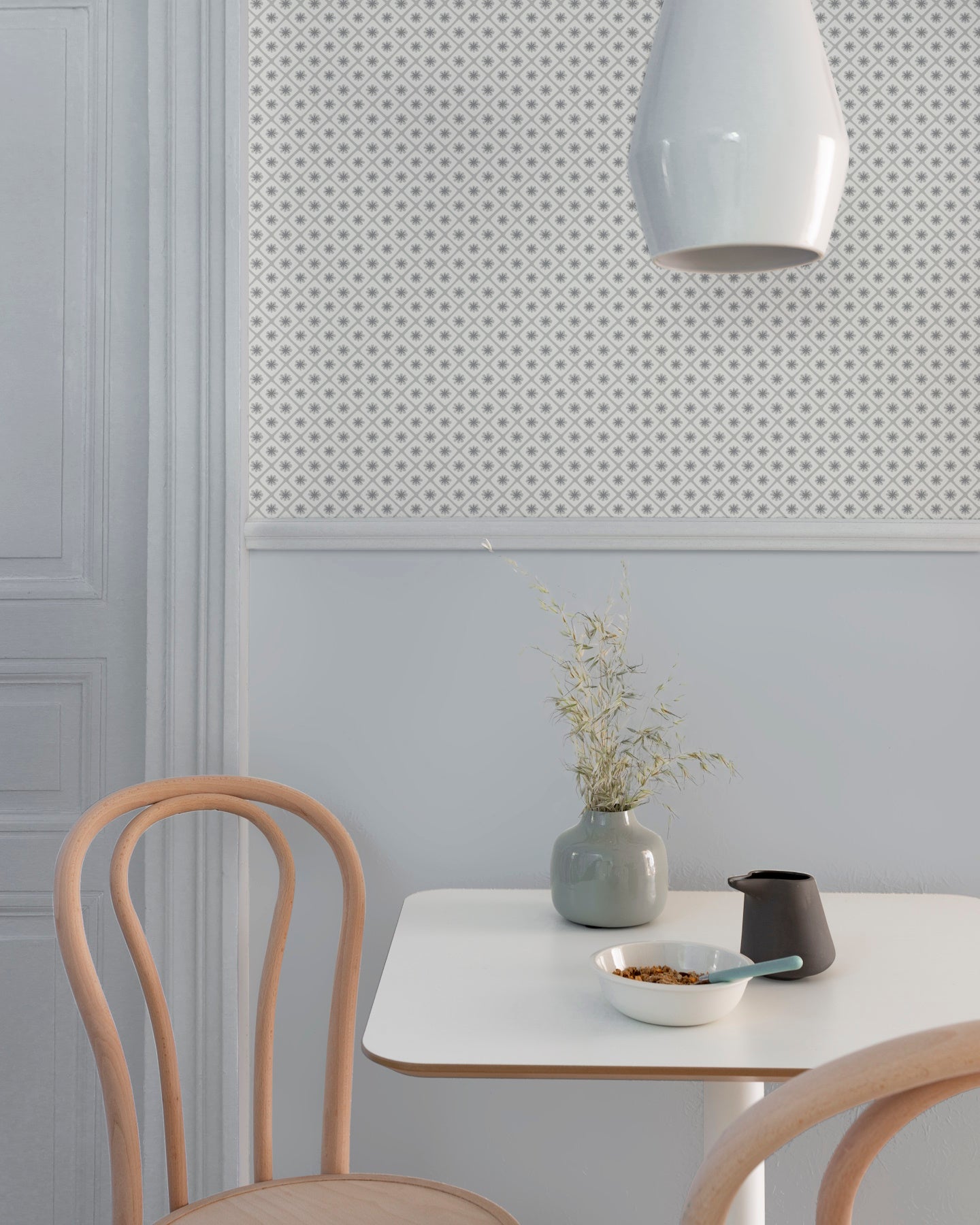 Coin repas aux murs bleu gris et motif géométrique Maître Domino Libéron, avec table blanche, chaises en bois clair et vase.
