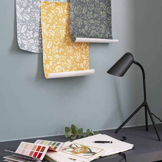 Mur bleu grisé, Velours de Peinture Libéron Oeillet de reine, avec lés fleuris, lampe noire et nuancier sur bureau.