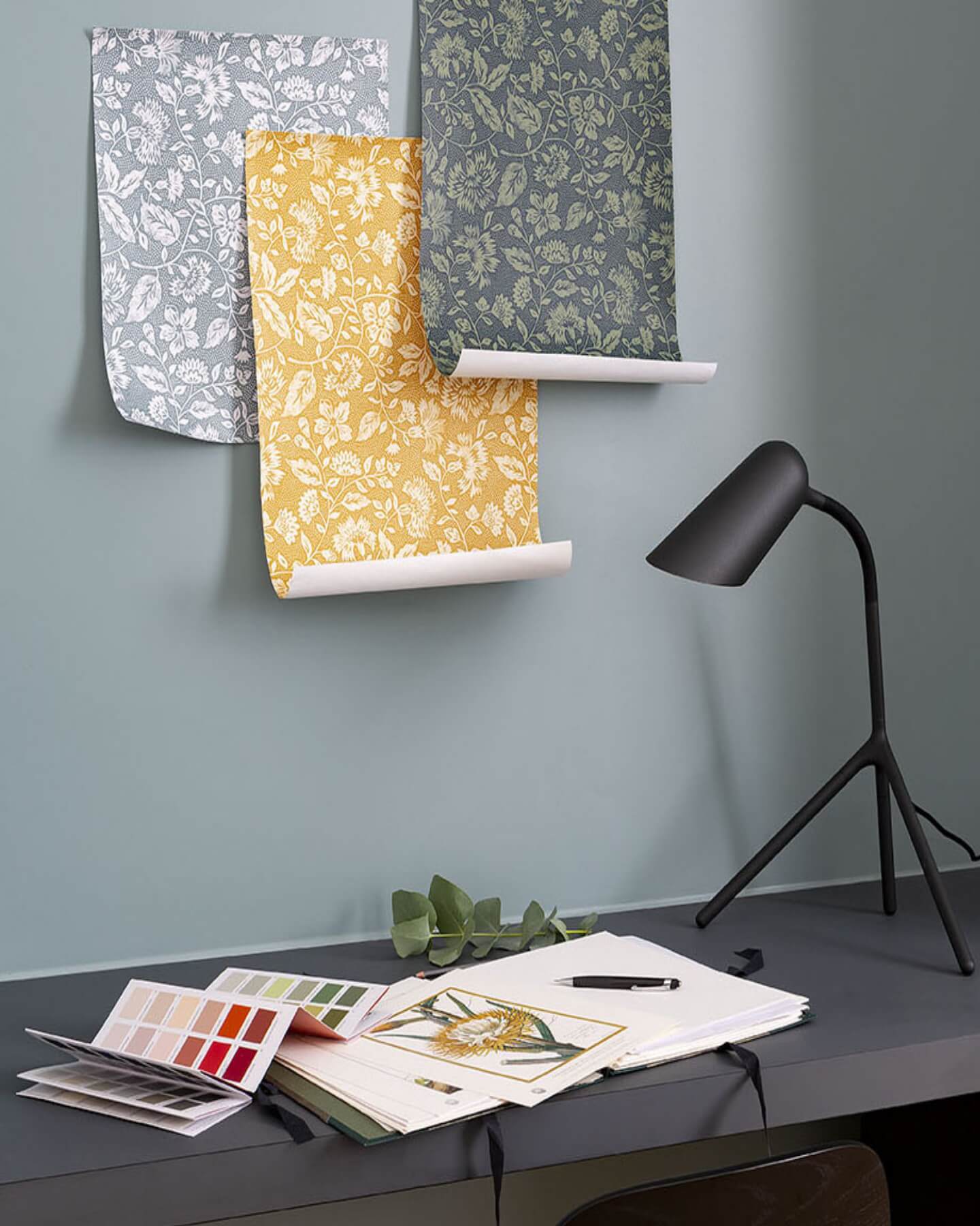Mur bleu grisé, Velours de Peinture Libéron Oeillet de reine, avec lés fleuris, lampe noire et nuancier sur bureau.