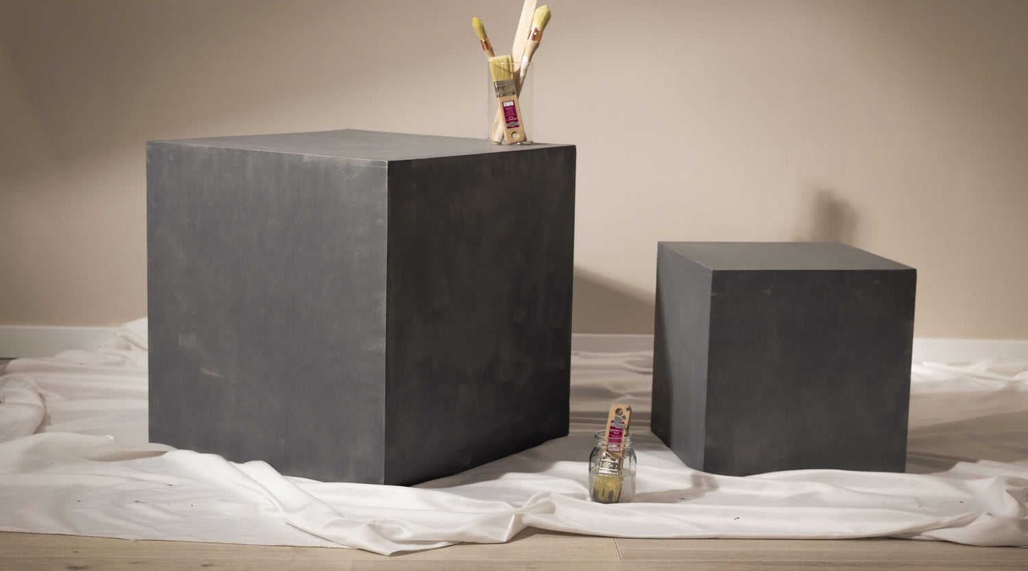 Cubes effet béton gris Libéron sur drap blanc, avec pinceaux et mur beige en arrière-plan.
