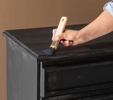 Application au pinceau d’une peinture effet patiné Libéron sur le plateau d’une commode noire.