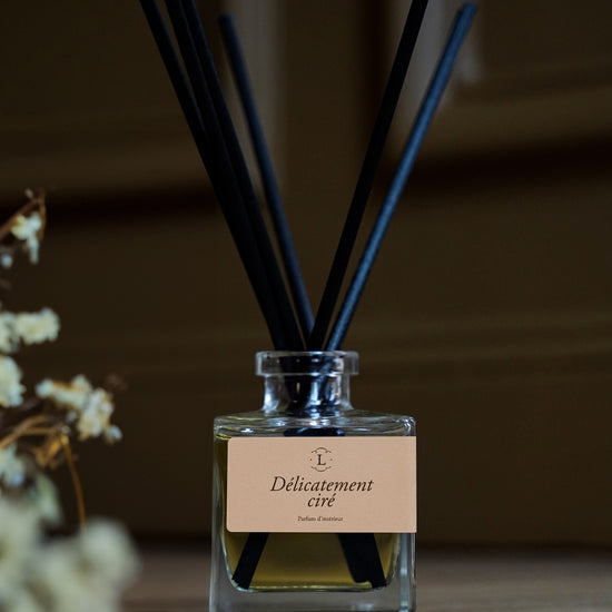 Diffuseur de parfum d’intérieur Libéron Délicatement ciré, flacon en verre avec tiges noires sur parquet.