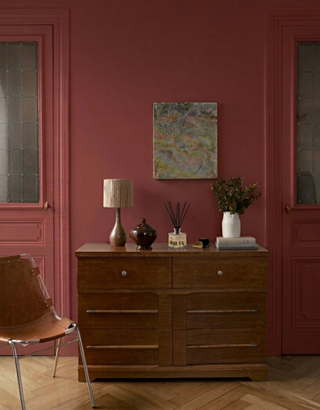 Salon avec une commode en bois foncé, une chaise en cuir marron, une petite lampe à poser et des murs peints en rouge et le parfum d'intérieur Libéron Délicatement Ciré.