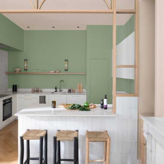 Cuisine vert impressionniste Libéron avec îlot blanc ambré, tabourets tressés et verrière en bois clair.