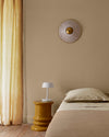 Chambre aux murs beige bottier Libéron, avec rideaux ocre, linge de lit beige et table d’appoint jaune moutarde.