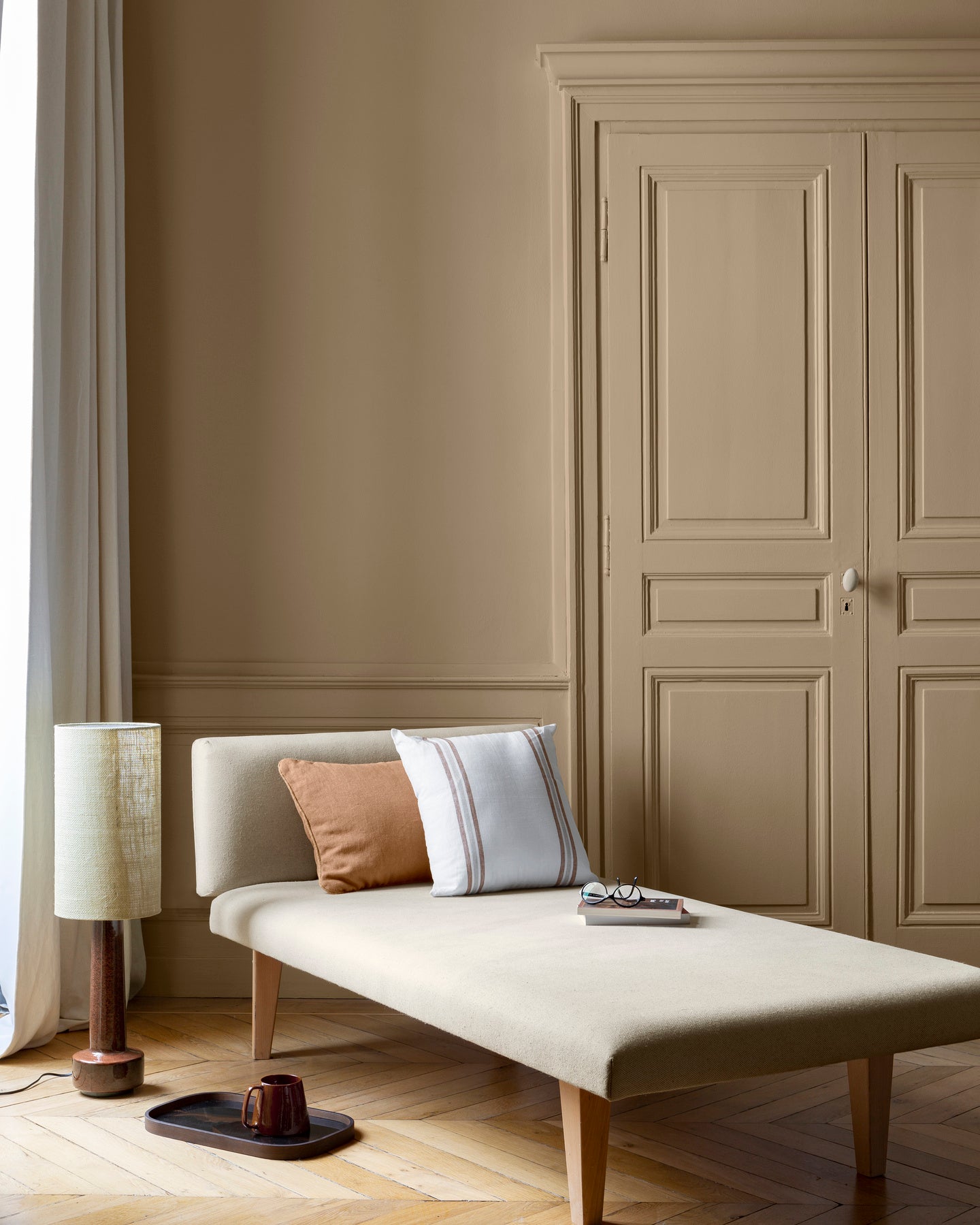 Salon aux murs beige bottier Libéron, avec méridienne écrue, portes assorties, coussins terracotta et parquet clair.
