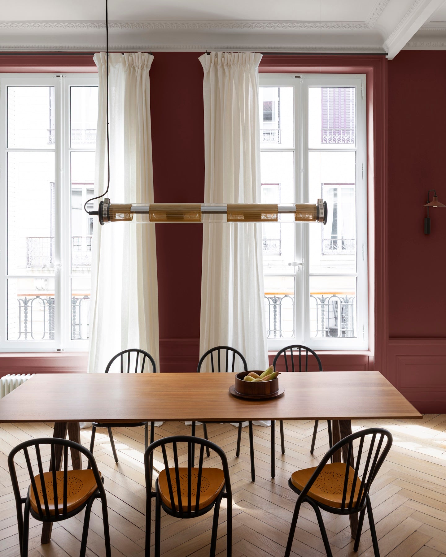 Salle à manger aux murs bordeaux tomette Libéron, avec grande table en bois, chaises noires et rideaux blancs.