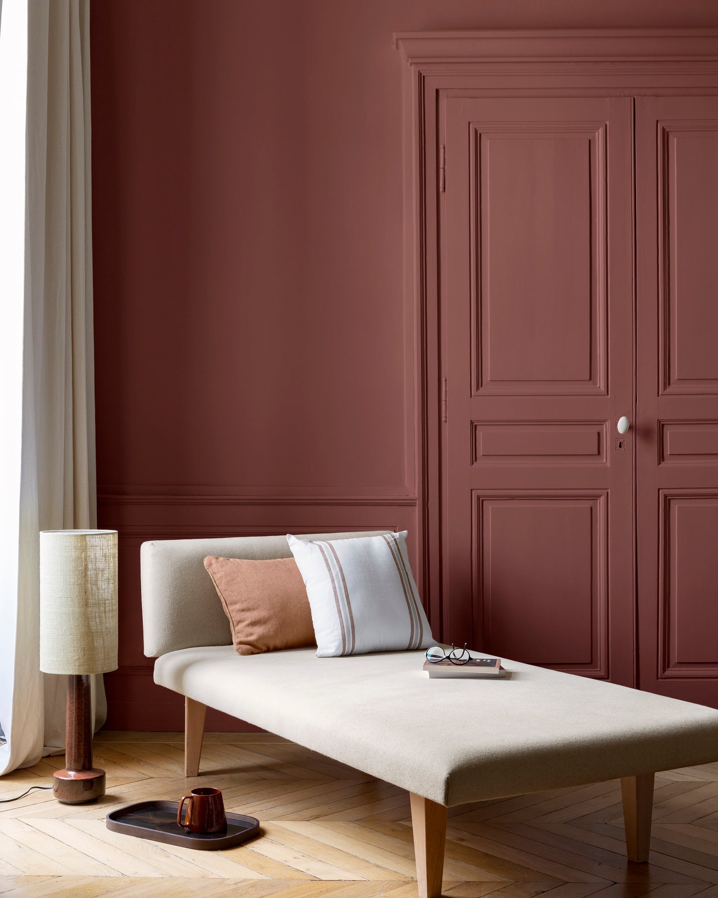 Salon aux murs bordeaux tomette Libéron, avec méridienne écrue, portes assorties, coussins terracotta et parquet clair.