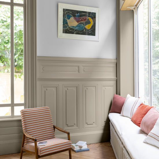Salon lumineux avec soubassement mouluré beiges, peinture 163 Faubourg Grège Galuchat Libéron, avec grandes fenêtres, fauteuil à rayures rouges et rebord de fenêtre utilisé comme canapé.