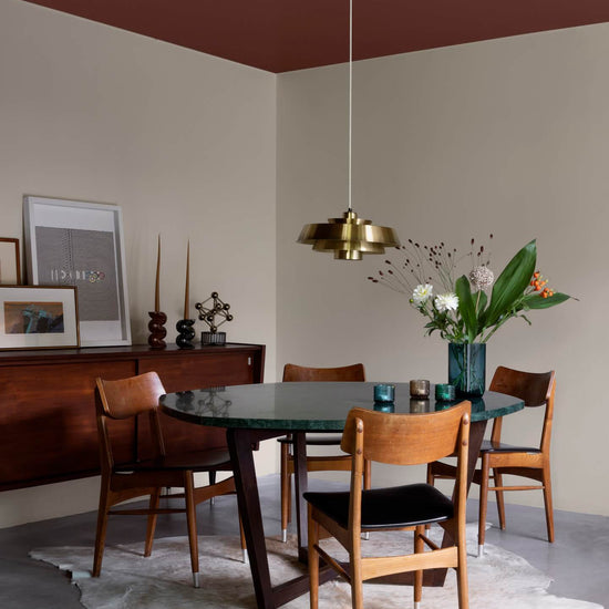 Salle à manger aux murs grège galuchat Libéron, avec table ronde sombre, chaises en bois et suspension dorée.