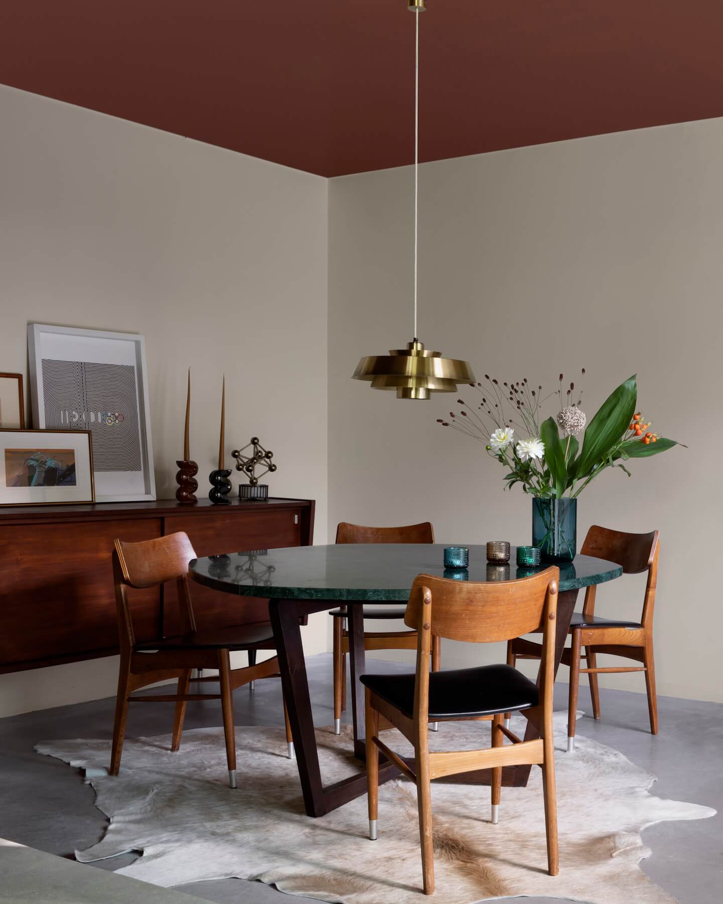Salle à manger aux murs grège galuchat Libéron, avec table ronde sombre, chaises en bois et suspension dorée.