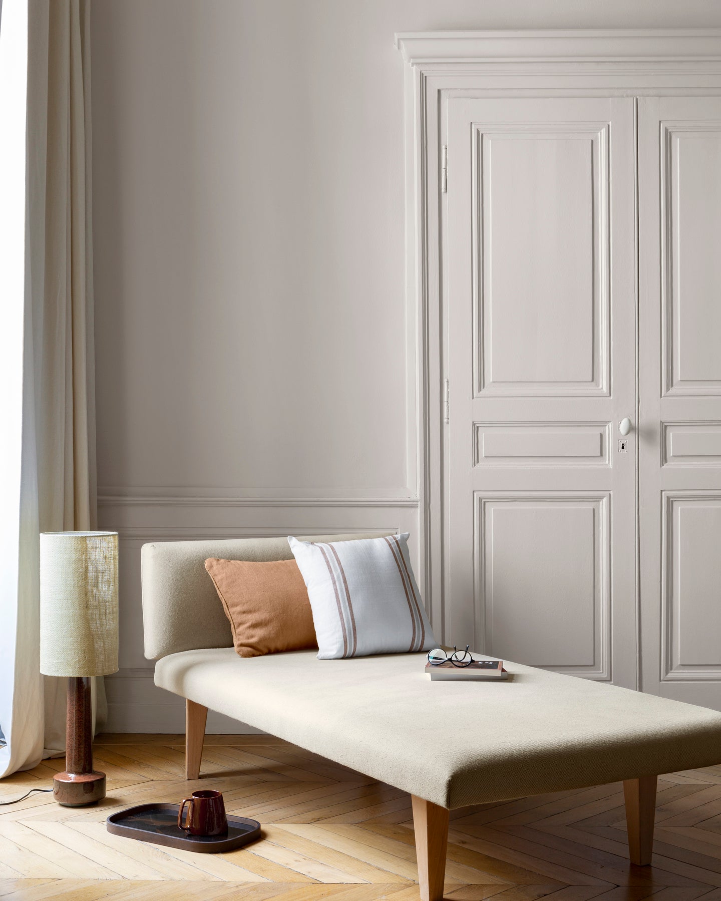 Salon aux murs grège tapissier Libéron, avec méridienne écrue, portes assorties, coussins terracotta et parquet clair.
