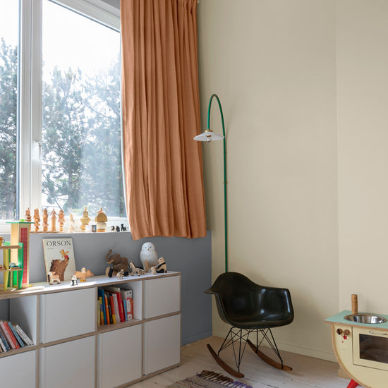 Chambre d’enfant aux murs ivoire orfèvre Libéron, avec rideaux terracotta, bibliothèque basse et fauteuil noir.