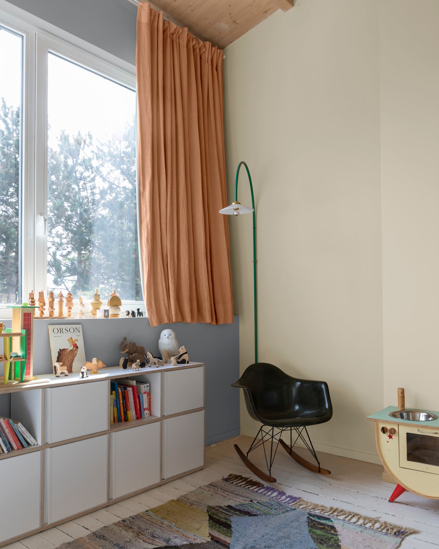 Chambre d’enfant aux murs ivoire orfèvre Libéron, avec rideaux terracotta, bibliothèque basse et fauteuil noir.