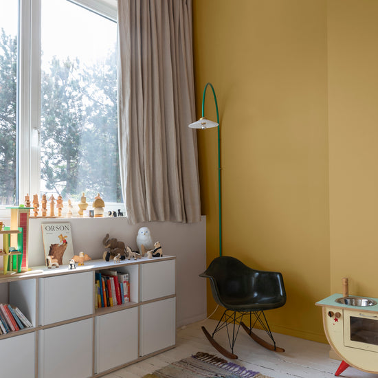Coin enfant aux murs jaune lumière Libéron, avec bibliothèque basse, fauteuil noir et grande fenêtre habillée de rideaux.