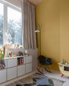 Coin enfant aux murs jaune lumière Libéron, avec bibliothèque basse, fauteuil noir et grande fenêtre habillée de rideaux.