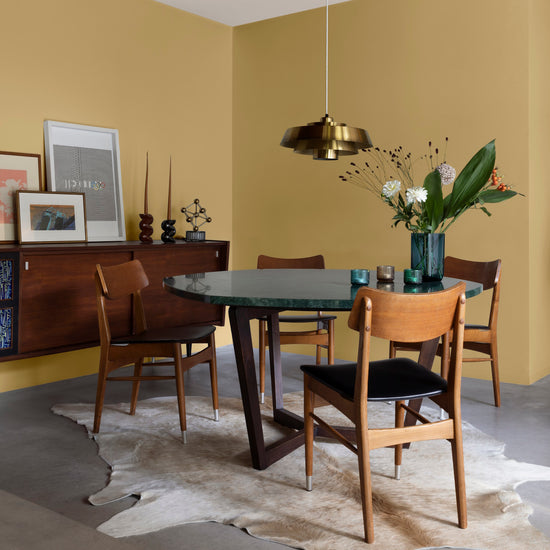 Salle à manger aux murs jaune lumière Libéron, avec table ronde, chaises en bois, buffet vintage et suspension dorée.