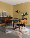 Salle à manger aux murs jaune lumière Libéron, avec table ronde, chaises en bois, buffet vintage et suspension dorée.