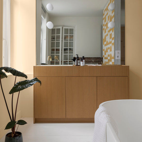 Salle de bain aux murs jaune tesselle Libéron, avec meuble vasque en bois clair, grand miroir et plante verte.