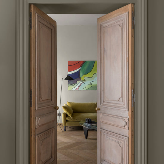 Double porte en bois ouvrant sur un salon, murs gris grande dame et lait d’amande Libéron, canapé vert et parquet.
