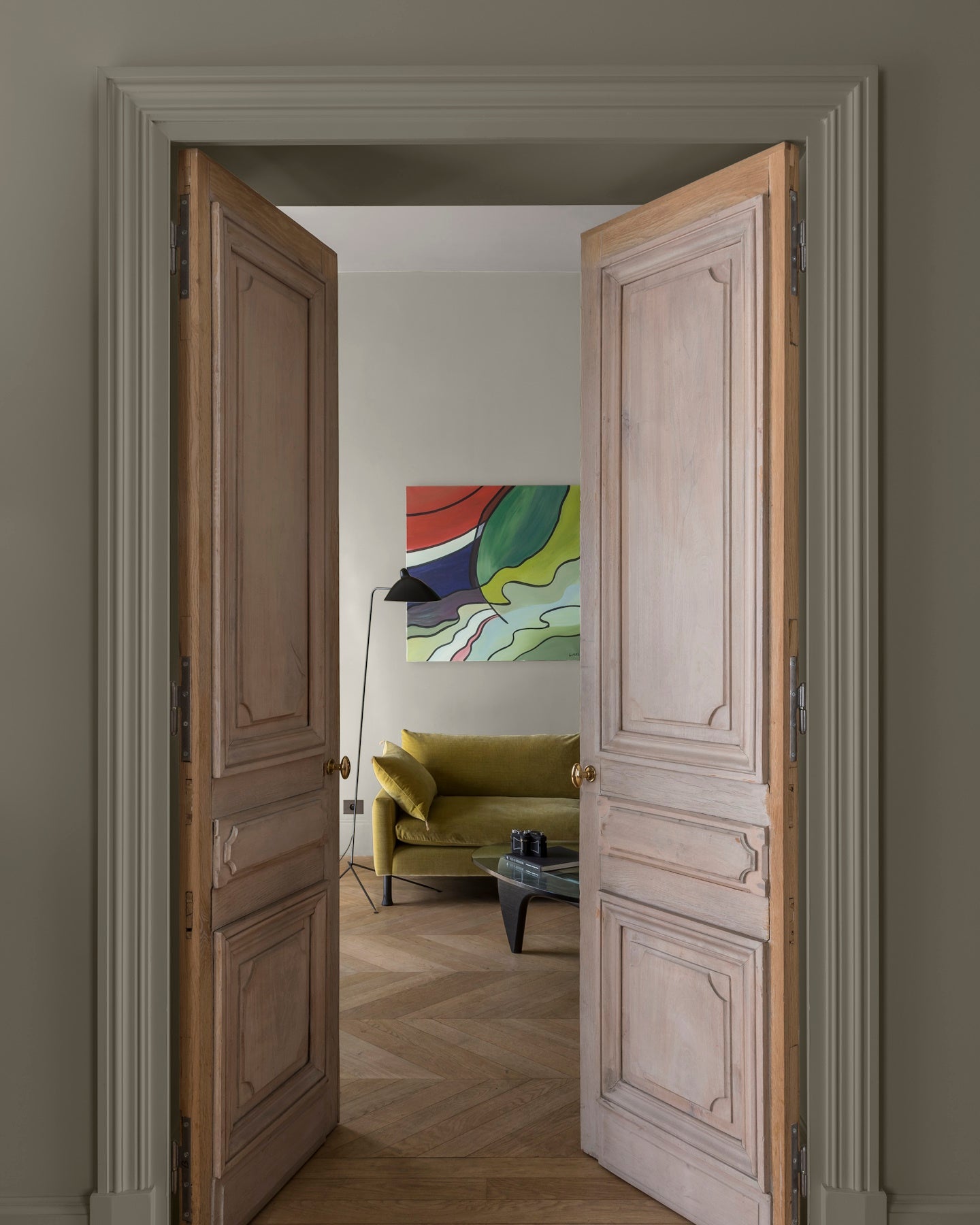 Double porte en bois ouvrant sur un salon, murs gris grande dame et lait d’amande Libéron, canapé vert et parquet.