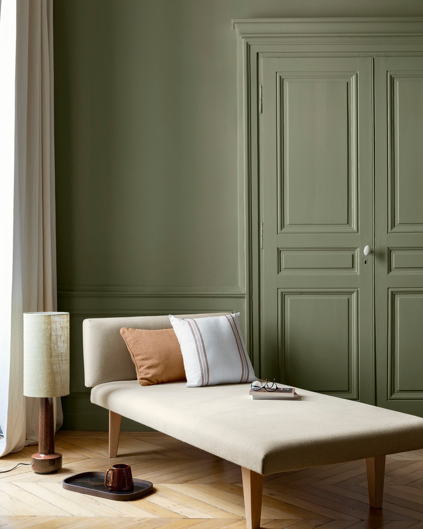 Salon aux murs Vert Ombrelle Libéron, avec méridienne écrue, portes assorties, coussins terracotta et parquet clair.