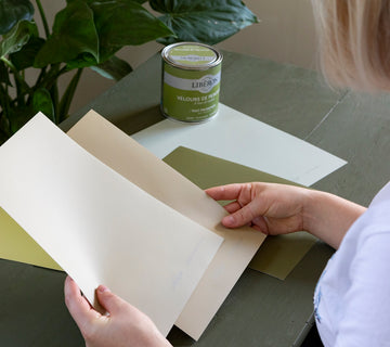 Femme tenant des échantillons de peinture Velours de Peinture, de couleurs beiges et verts, pot de Velours de Peinture Vert Herbarium en arrière-plan.