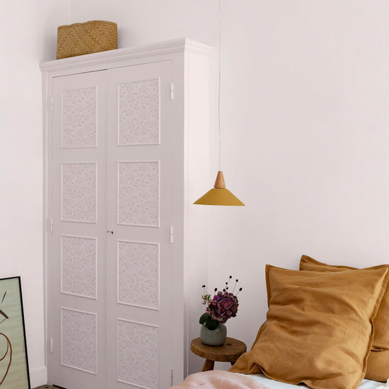 Chambre aux murs rose poudré, avec armoire blanche à panneaux floraux Belle Jeanne Libéron, suspension ocre et linge de lit rose.
