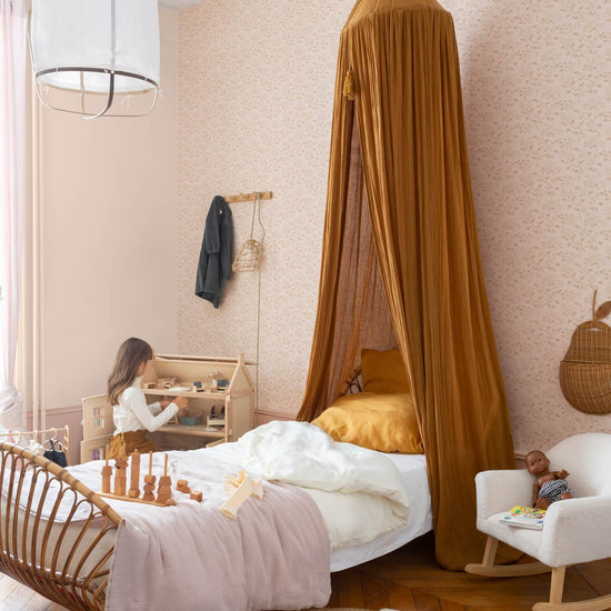 Chambre d’enfant au mur floral rose poudré, teinte Belle Jeanne Libéron, avec lit en rotin, baldaquin ocre et fauteuil blanc.
