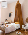 Chambre d’enfant au mur floral rose poudré, teinte Belle Jeanne Libéron, avec lit en rotin, baldaquin ocre et fauteuil blanc.