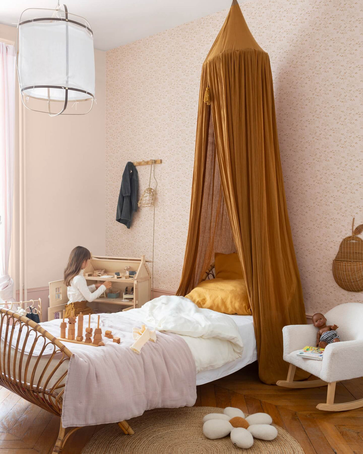 Chambre d’enfant au mur floral rose poudré, teinte Belle Jeanne Libéron, avec lit en rotin, baldaquin ocre et fauteuil blanc.