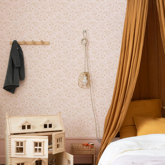 Détail de chambre d’enfant au mur floral rose poudré, teinte Belle Jeanne Libéron, avec lit, baldaquin ocre et maison de poupée.