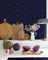 Cuisine au mur floral bleu nuit, teinte Belle Jeanne Libéron, avec plan de travail blanc, lampes et légumes violets.