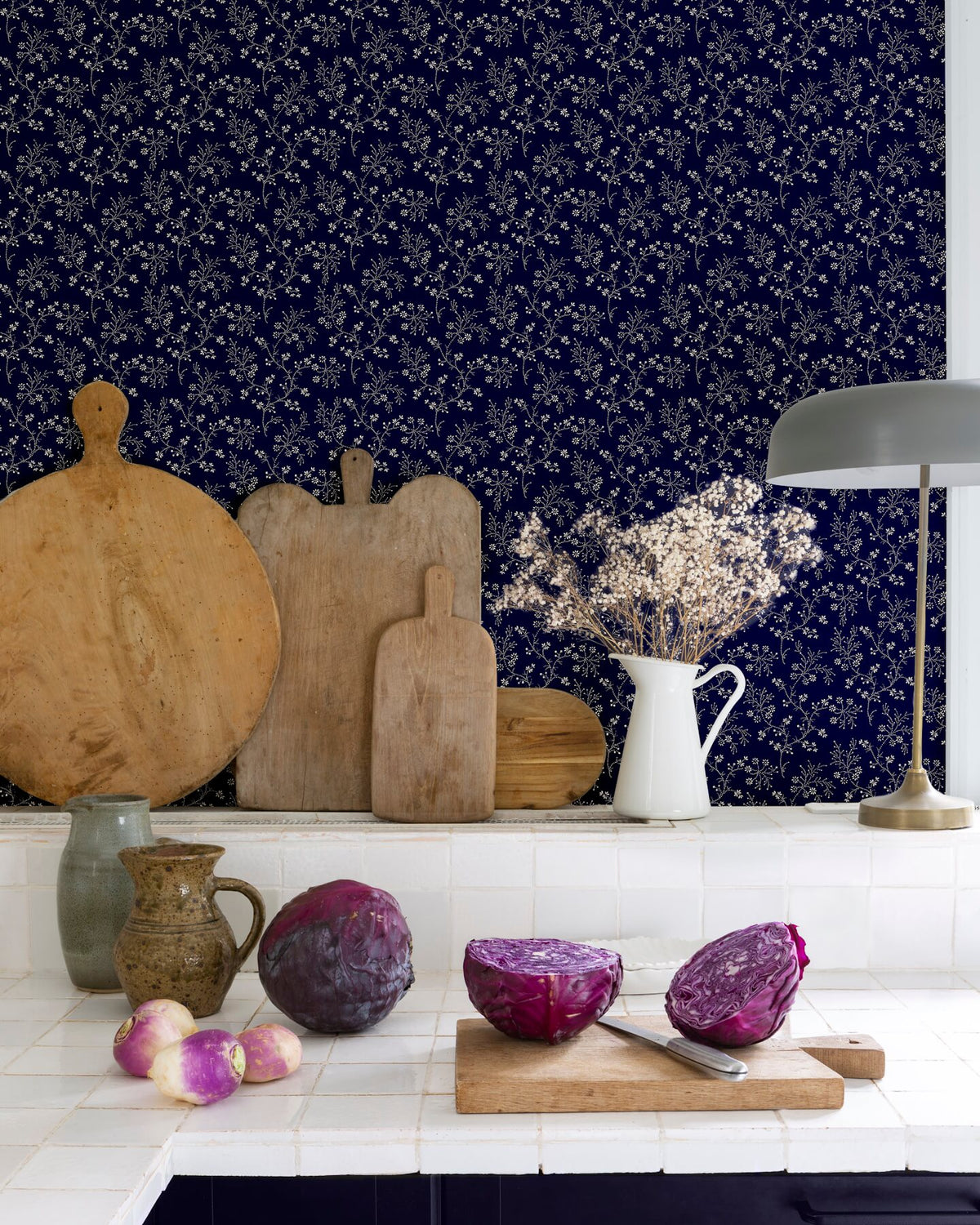 Cuisine au mur floral bleu nuit, teinte Belle Jeanne Libéron, avec plan de travail blanc, lampes et légumes violets.