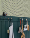 Coin cuisine avec papier peint Velours de Papier Florette n°3 Libéron, soubassement vert foncé, tablier et outils de nettoyage suspendus sur des accroches murales.
