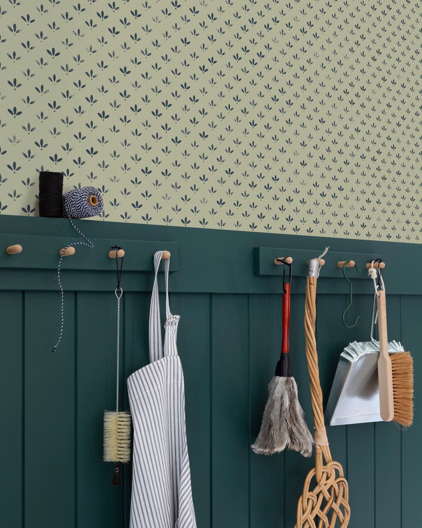 Coin cuisine avec papier peint Velours de Papier Florette n°3 Libéron, soubassement vert foncé, tablier et outils de nettoyage suspendus sur des accroches murales.