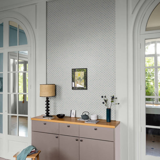 Entrée lumineuse au mur Florette Libéron, avec papier peint blanc à petit motif bleu, meuble taupe et portes vitrées blanches.