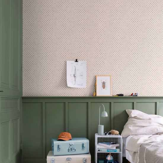 Chambre aux murs Florette Libéron, avec papier peint crème à petit motif vert, soubassement vert sauge, lit clair et valises pastel.