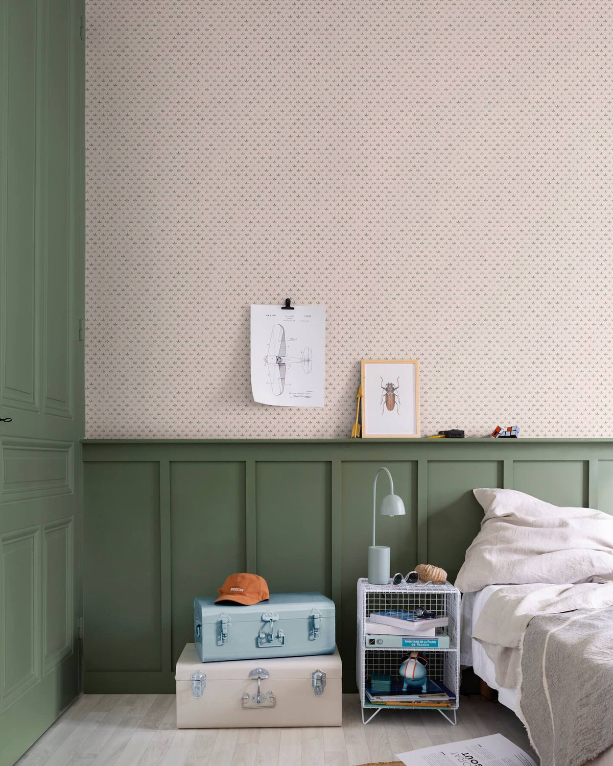 Chambre aux murs Florette Libéron, avec papier peint crème à petit motif vert, soubassement vert sauge, lit clair et valises pastel.