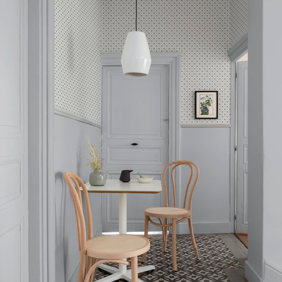 Coin repas aux murs gris perle et papier peint Maître Domino Libéron, avec table blanche, chaises en bois et suspension blanche.